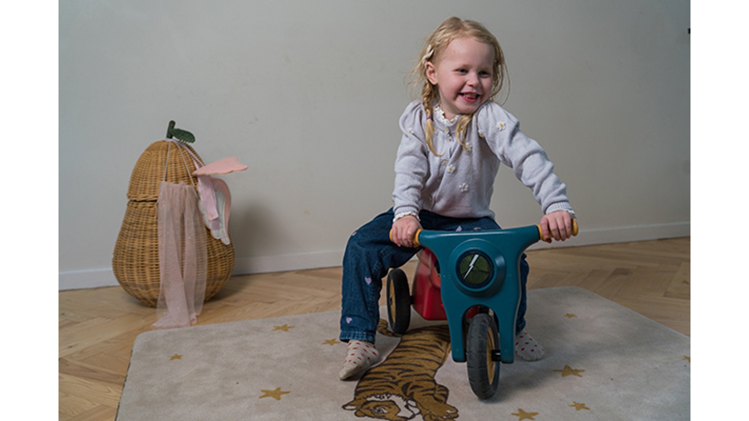 Et barn på en lyseblå motorcykel-gynge med lyserødt legetøj og flettet kurv på trægulv.