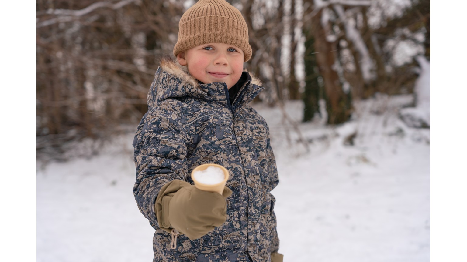 Person i vinterbeklædning holder en snebold i en snedækket skov.