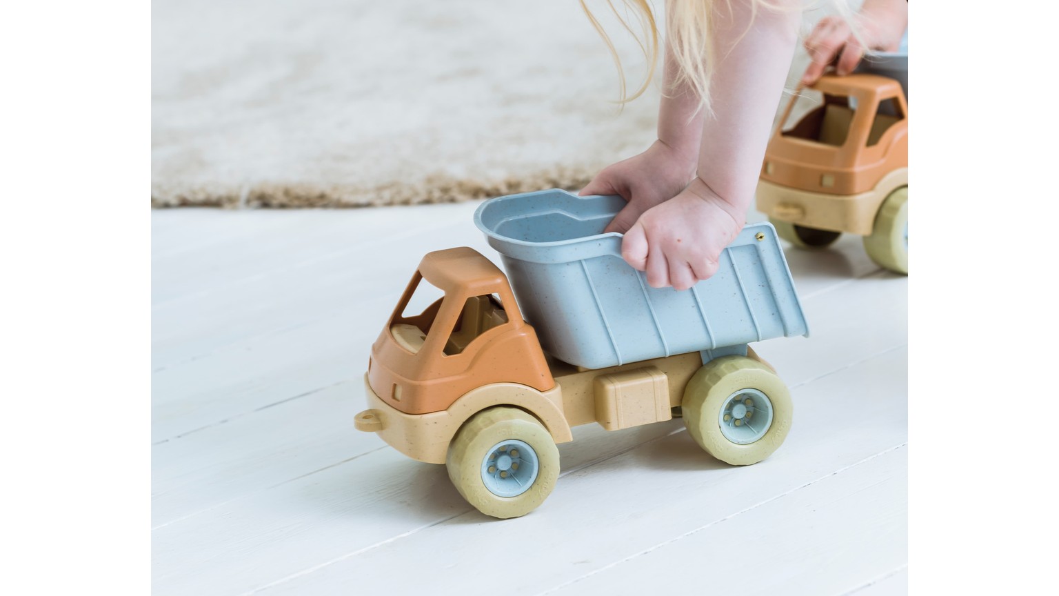 Legetøjslastbil i beige med blå last på hvidt underlag. Lille fod træder på lasten.