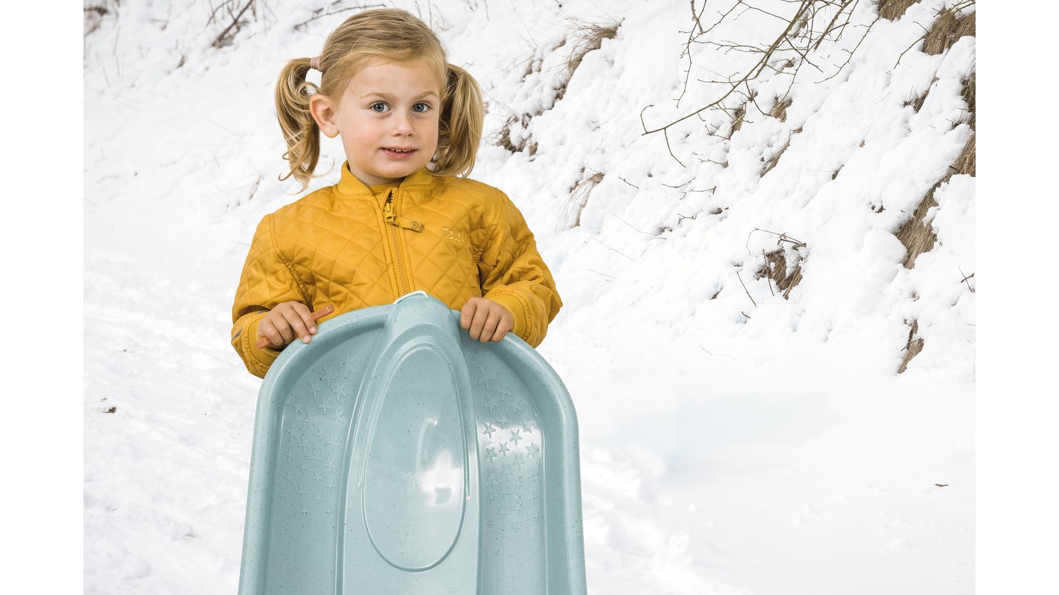 Barn i gul trøje på lyseblå bobslæde med snedækket baggrund.