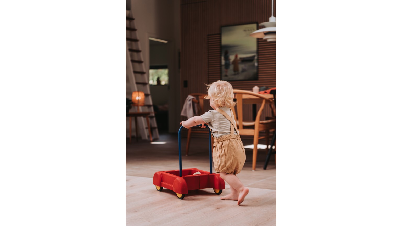 Lille barn trækker rød trækvogn på hjul henover et trægulv i et hjemligt miljø
